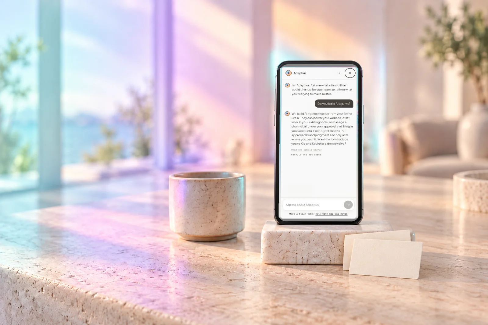 A phone on a stone stand showing the adaptius.ai assistant answering a question, beside a ceramic cup and two blank brand rule cards, in coloured daylight.
